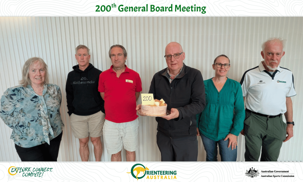 Six adults pose indoors, holding a cake with '200', celebrating the 200nd General Board Meeting.