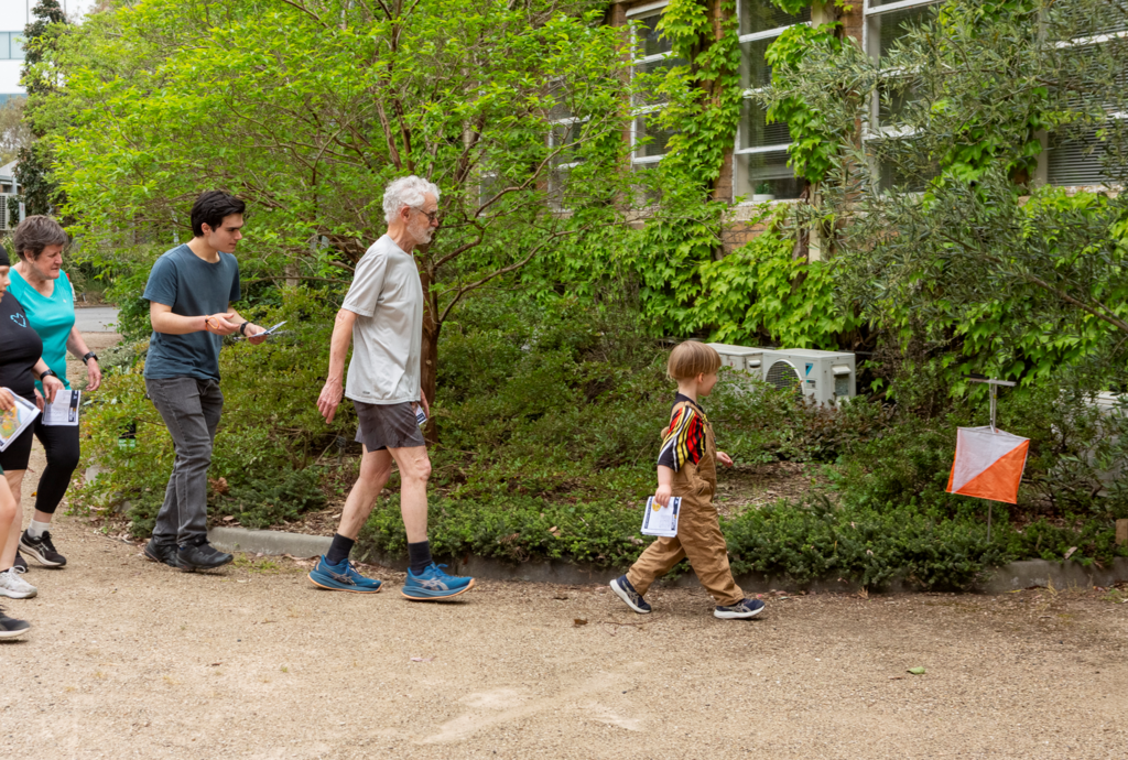 Club_event_hero - Orienteering Australia - Orienteering Australia Group of adults and a child in a brown costume walking along a garden path, holding papers; orange-and-white flag marker to the right.