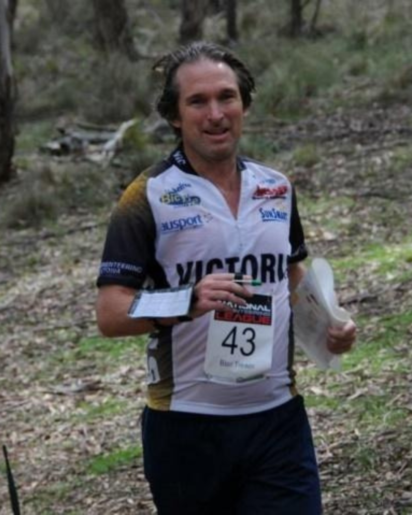 Male trail runner in a white and black race jersey with number 43, moving through a wooded trail, holding a paper or map and a plate in his hands.