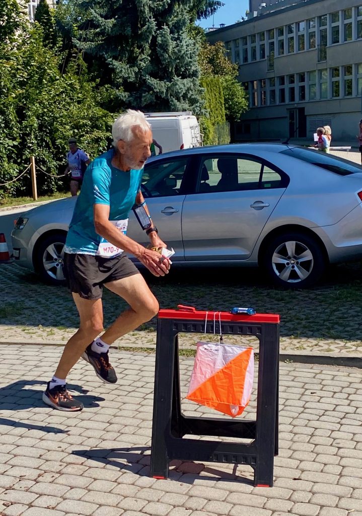 IMG_1148 - Orienteering Australia An elderly man with white hair, wearing a turquoise shirt and black shorts, runs on a cobblestone street during a race with a bib number visible on his waist.