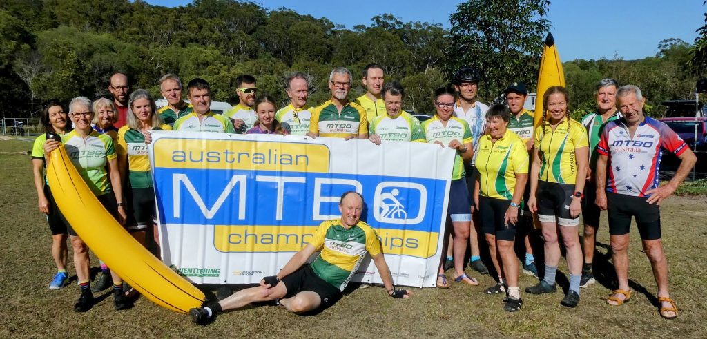 Group of cyclists in green/yellow uniforms posing outdoors with a large banner reading 'Australian MTB Championships'.