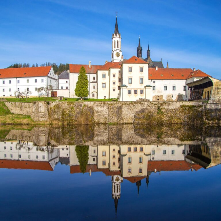 Historic European riverside with white buildings, red roofs, and church spires, reflected in calm water