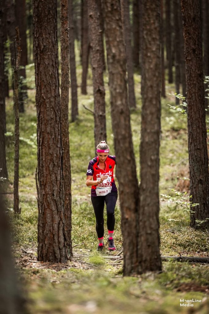 image5 - Orienteering Australia Female trail runner in a red athletic top and black tights, wearing a pink headband, running through a pine forest with a race bib showing number 55532.