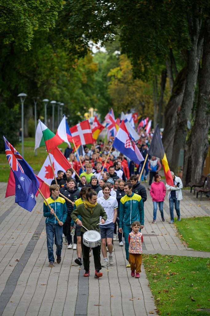 820114527_1798999018896527_5623262040768707954_n - Orienteering Australia Crowd of people marching down a park path with international flags, including Australia, Canada, and the UK, lined by trees and lampposts.