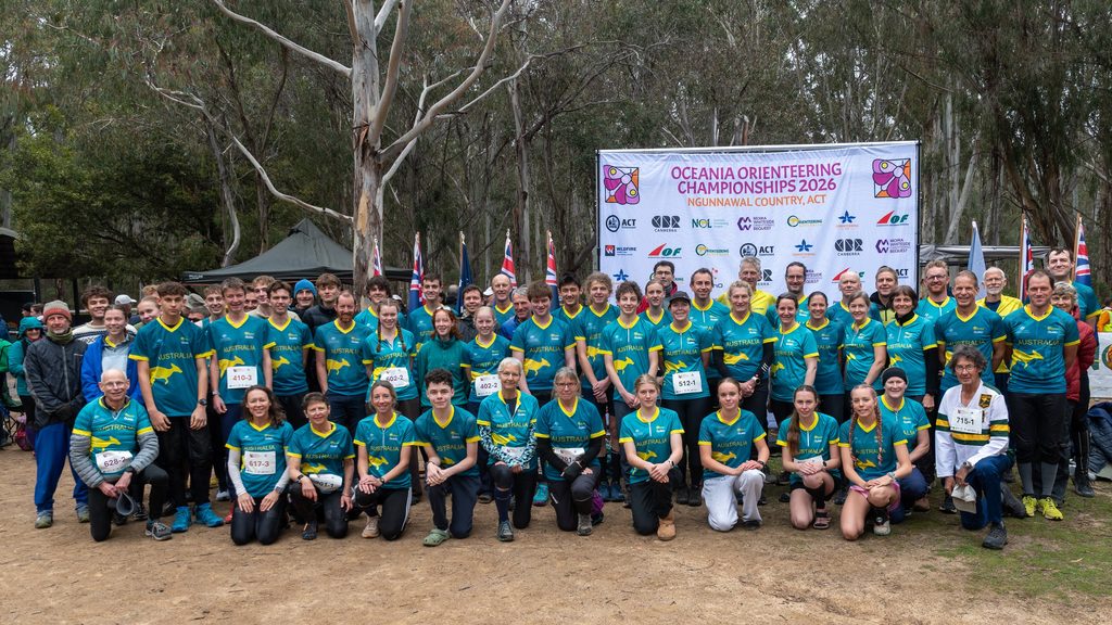 Large group of athletes in teal Australia jerseys posing for a team photo at the Oceania Orienteering Championships 2026, with a banner and flags in the background.