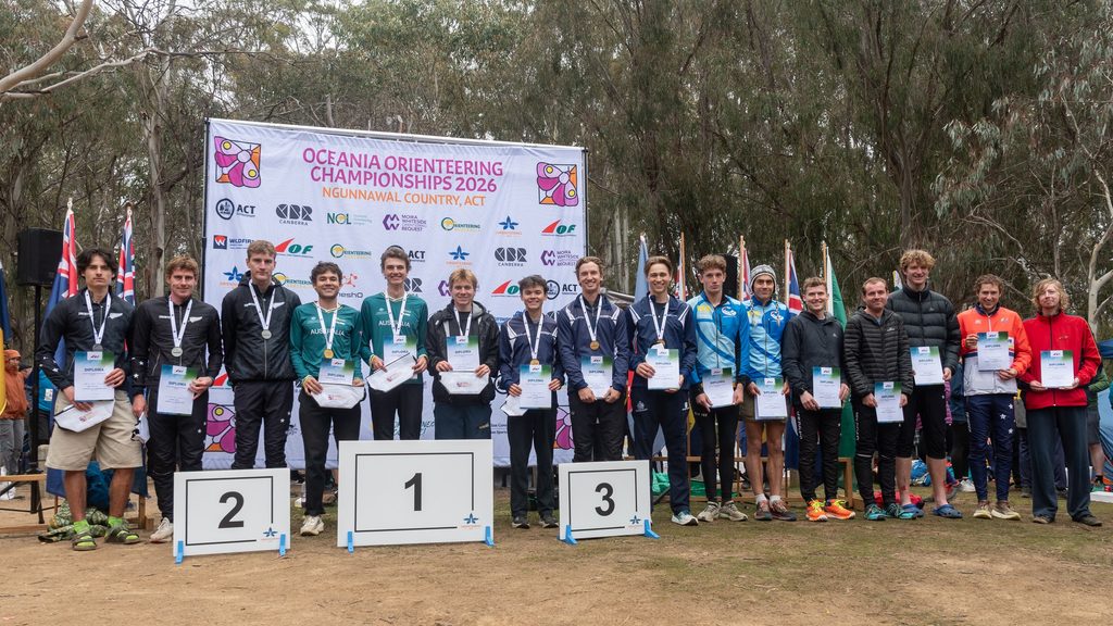 Young athletes stand on a podium holding diplomas and medals after an awards ceremony, with a banner reading 'Oceania Orienteering Championships 2026' behind them.
