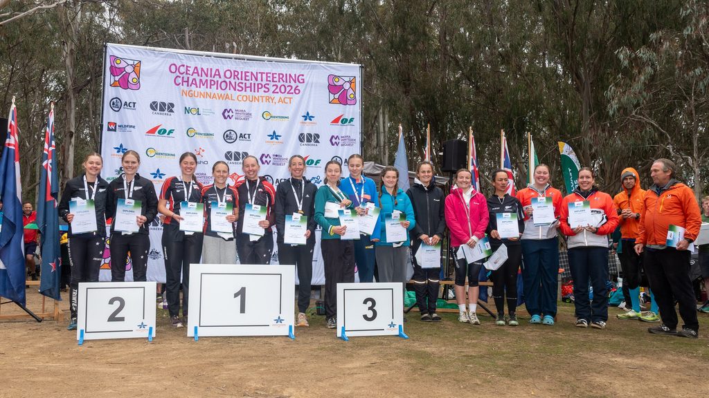 Athletes stand on a podium holding diplomas at the Oceania Orienteering Championships 2026, with a banner and flags in the background.