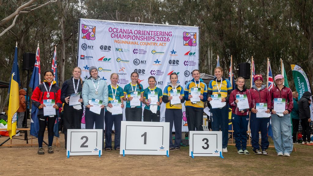 Athletes on a podium with diplomas and medals at the Oceania Orienteering Championships 2026 award ceremony, banners and flags in the background