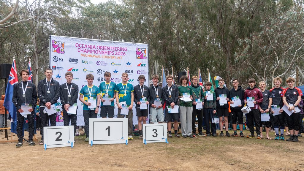 Group of young athletes on a podium holding diplomas at the Oceania Orienteering Championships 2026 awards ceremony under a banner.