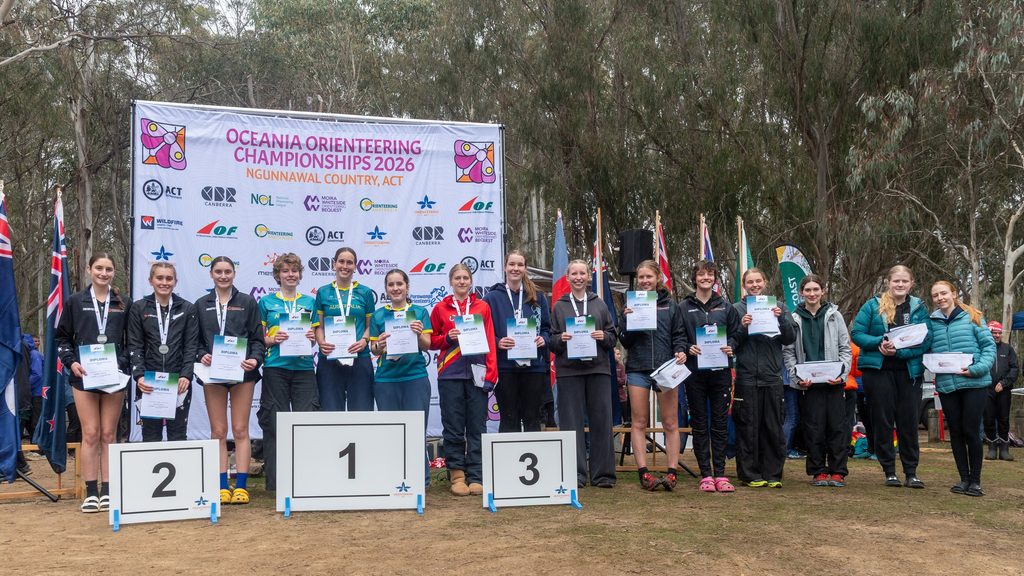 Group of young athletes on podiums holding diplomas at the Oceania Orienteering Championships 2026 ceremony in front of a banner and flags.