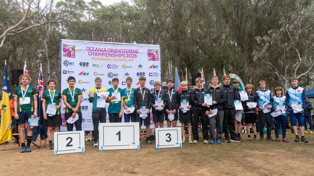 Group of young athletes standing on podiums with medals at the Oceania Orienteering Championships 2026, holding certificates, in front of a banner and trees.