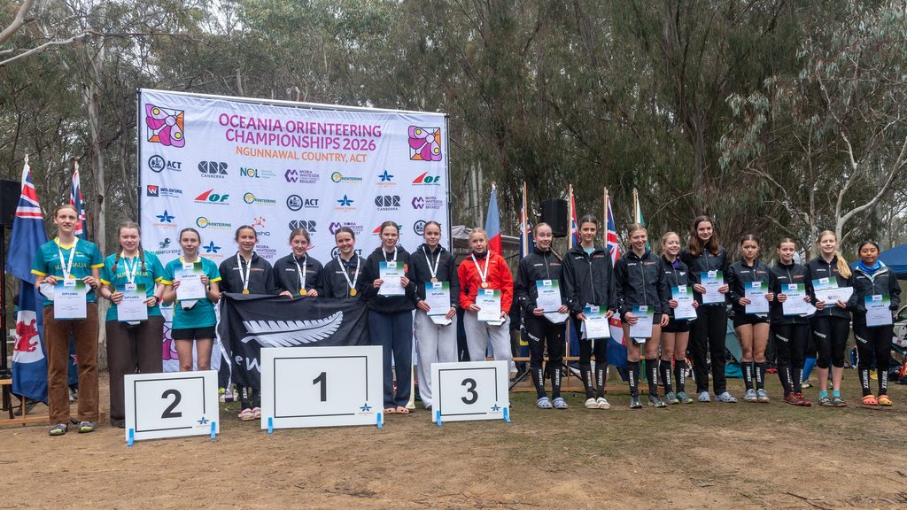 Young competitors on a podium at the Oceania Orienteering Championships 2026 awards, holding diplomas and medals with flags behind them.