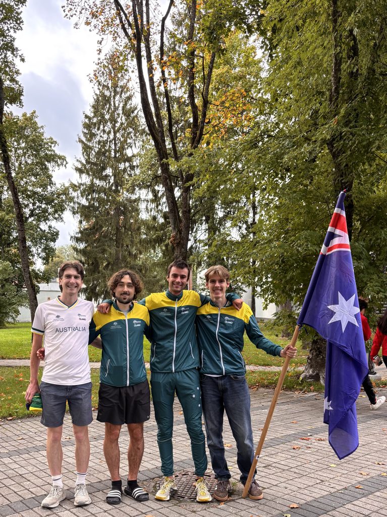 Photo 22-9-2026 4 58 29 pm - Orienteering Australia Group of four young men standing outdoors with arms around each other, wearing athletic jackets; one holds a blue flag with stars. Autumn trees in background.