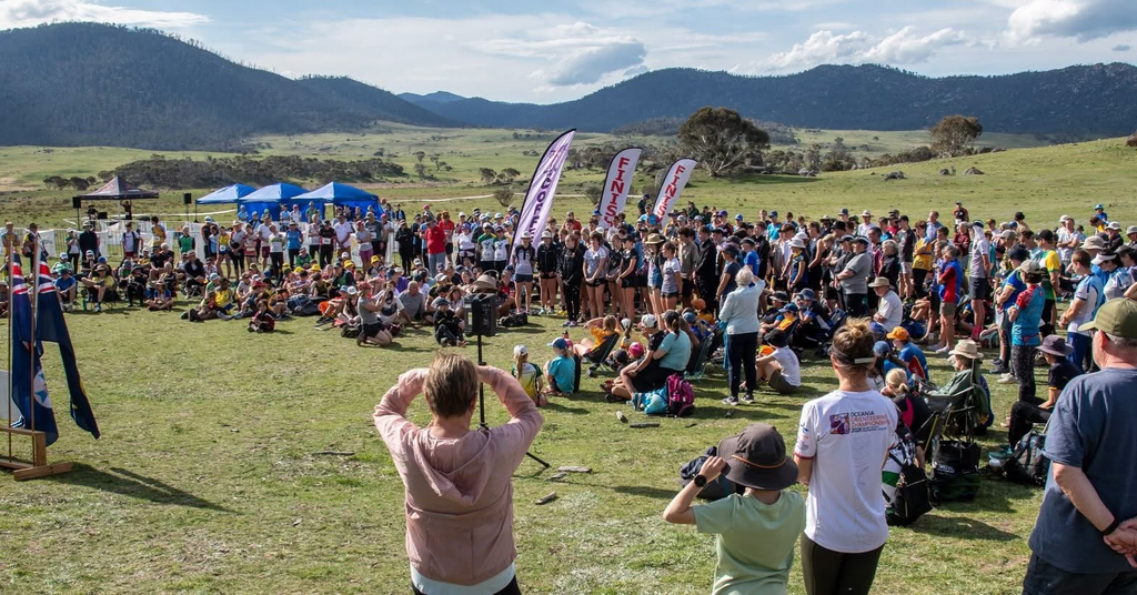 Large crowd gathered on an open field for an outdoor event with tents, flags, and mountains in the background.