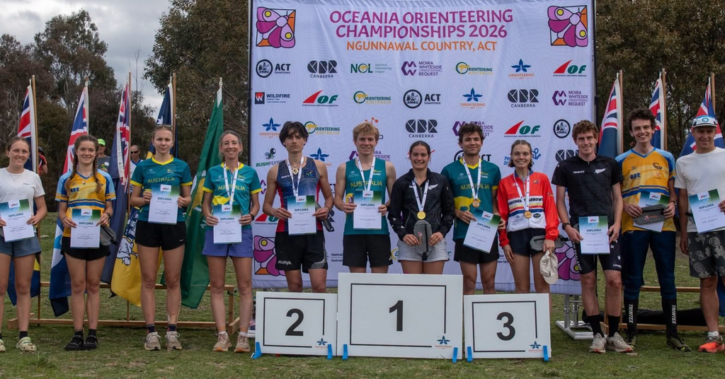 Group of young athletes standing on a winners' podium with diplomas at the Oceania Orienteering Championships 2026 backdrop, flags behind them.