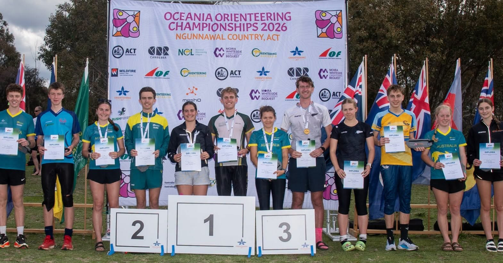 Group of young athletes on a winners' podium holding diplomas at the Oceania Orienteering Championships 2026 ceremony with flags and a banner backdrop.