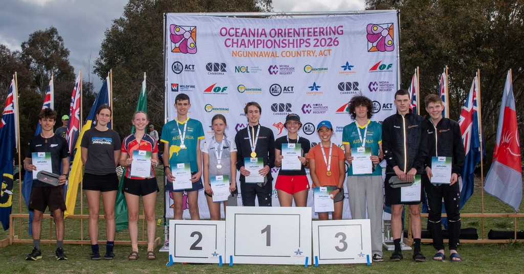 Group of young athletes on a podium holding diplomas and medals at the Oceania Orienteering Championships 2026 stage backdrop with flags nearby
