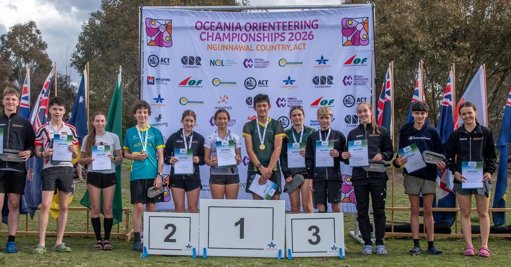 Group of young athletes on a podium at the Oceania Orienteering Championships 2026, holding diplomas and medals with a banner behind them and flags on both sides.