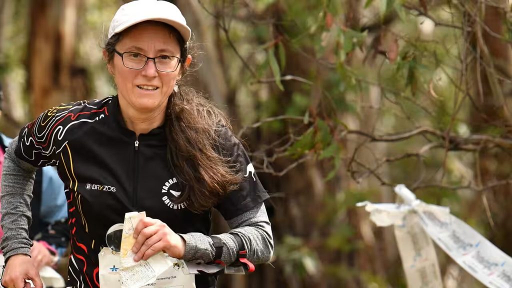 image0 2 - Orienteering Australia Woman runner in a black sports shirt and white cap, holding a race bib and papers outdoors in a wooded area.