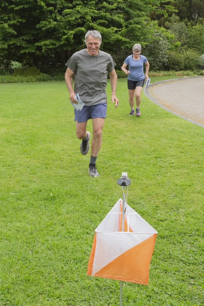 Two runners on a grassy park path: a man in a gray shirt leads ahead of a woman, with an orange-and-white course marker in the foreground.