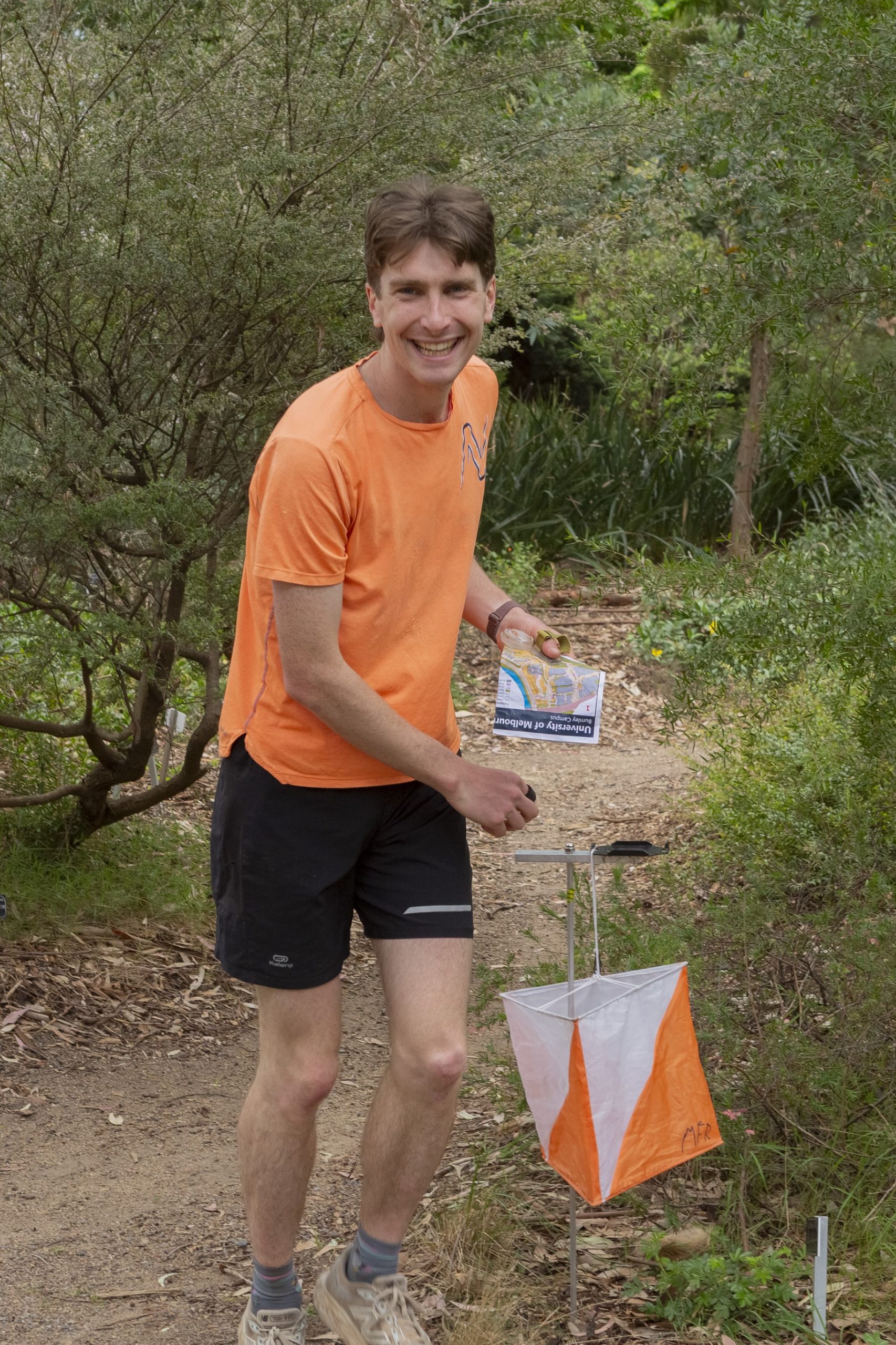 Smiling man in an orange shirt holds a map while standing on a dirt path with orange-white orienteering marker flags nearby in a green, bushy outdoor area.