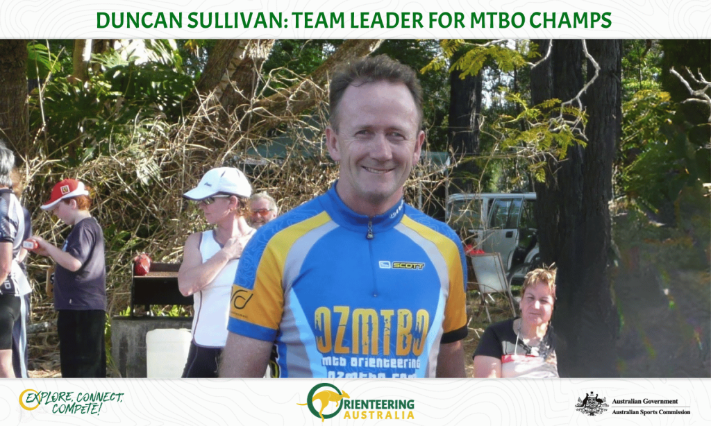 Smiling man in blue and yellow cycling jersey at a park team event, with spectators in the background and banners visible.