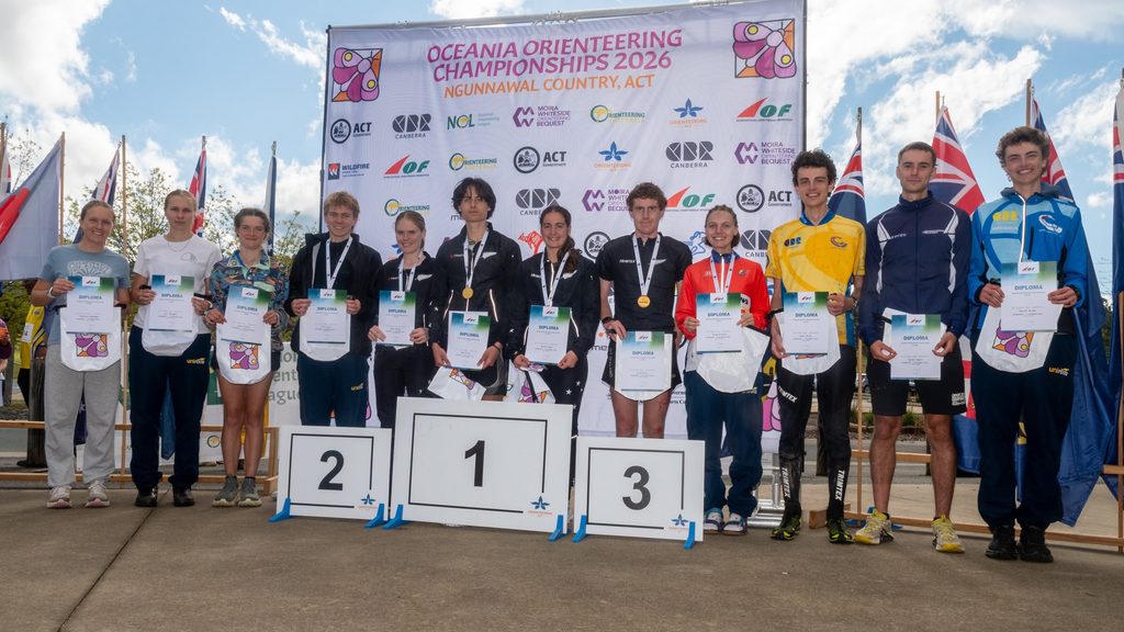 Group of young athletes on a podium holding diplomas, medals, and awards at the Oceania Orienteering Championships 2026 ceremony, with a large banner in the background and flags on either side.