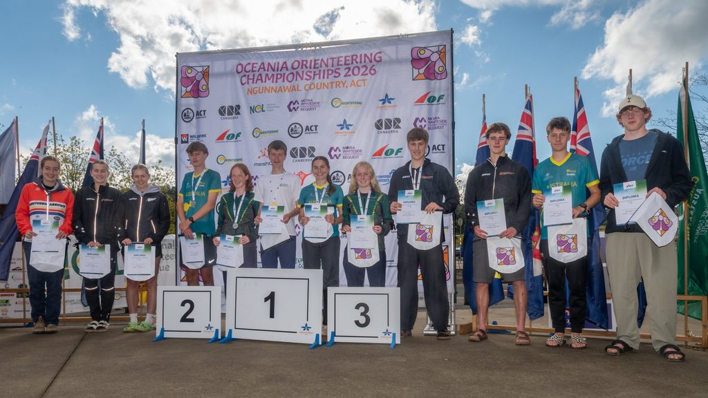 Group of athletes on a three-step podium holding diplomas at the Oceania Orienteering Championships 2026 stage, with flags and a branded backdrop behind them.