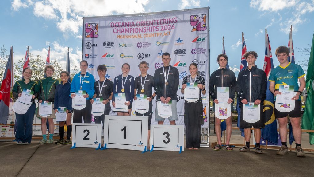 Group of young athletes on a winners' podium holding diplomas at the Oceania Orienteering Championships 2026, with a banner and flags behind them.