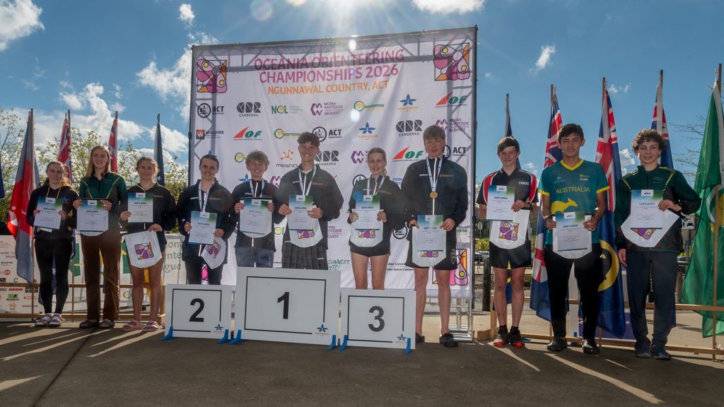 Young competitors pose on podiums with diplomas at an awards ceremony for the Oceania Orienteering Championships 2026.