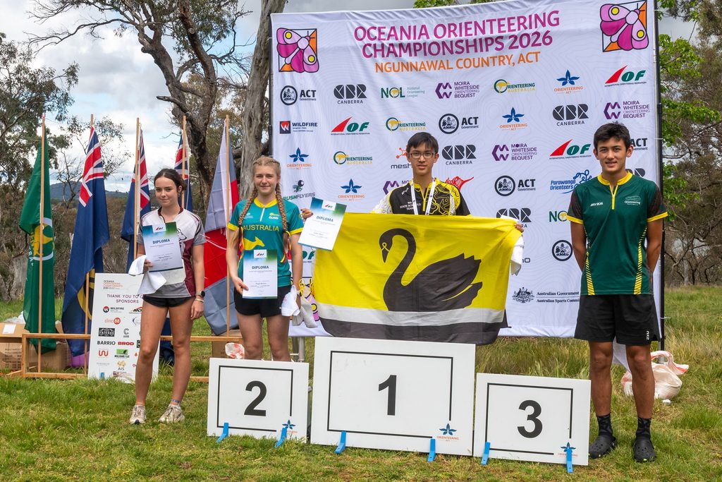 Podium scene at the Oceania Orienteering Championships 2026 with three young competitors holding diplomas and a large event banner behind them.