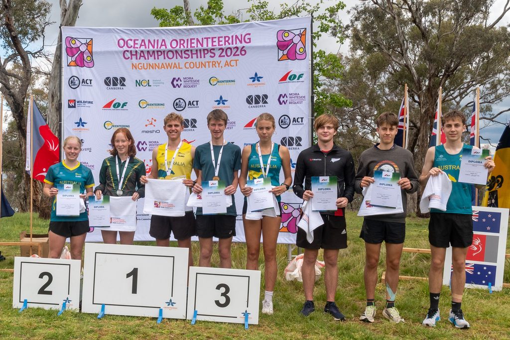 Group of young athletes on a winners' podium holding diplomas at the Oceania Orienteering Championships 2026 banner behind them.