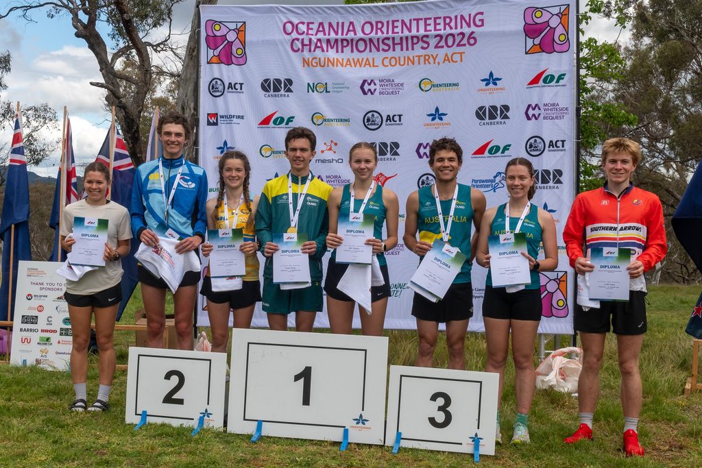 Group of young athletes on a podium holding diplomas and medals at the Oceania Orienteering Championships 2026 banner behind them.
