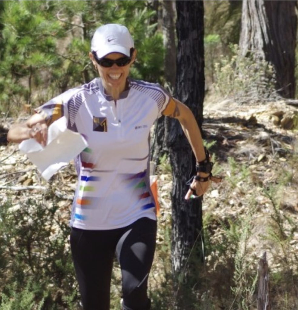 Female trail runner on a forest path, holding a folded map and smiling, wearing a white cap and sunglasses.